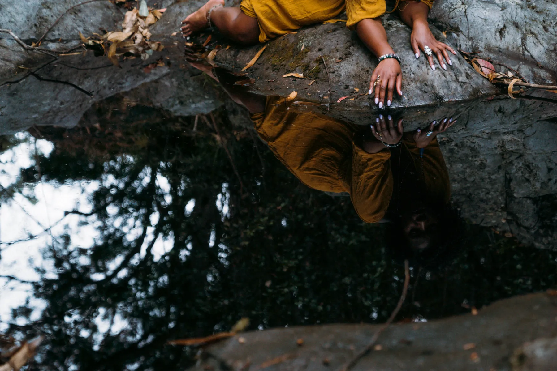 A woman resting on a rock, her reflection visible in still water below
